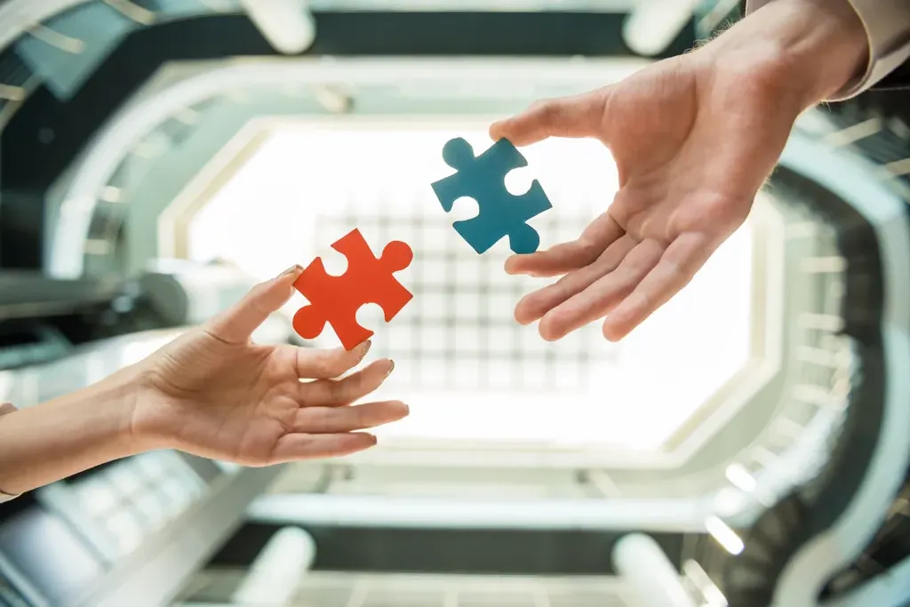Two hands each holding a puzzle piece, one red and one blue, ready to assemble, with a blurred modern building in the background.